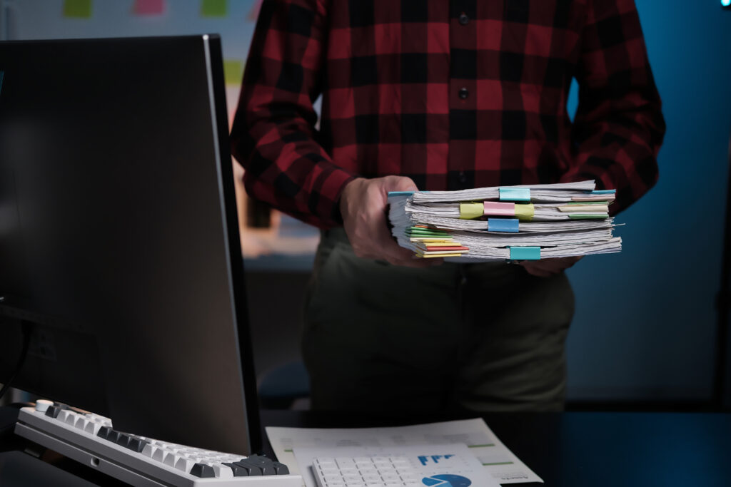A man is holding a stack of papers in front of a computer monitor. The papers are organized and labeled with different colors