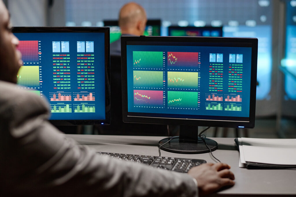 Rear view of modern African American broker sitting in front of computer monitors watching and analyzing previous stock trading stats in evening