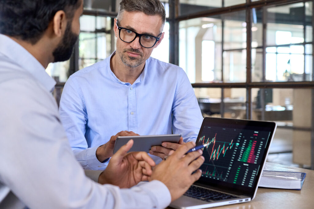 Diverse managers traders analysts discussing financial growth market at desk with laptop with graphs on screen using tablet device. Investors brokers analysing indexes online cryptocurrency stock.
