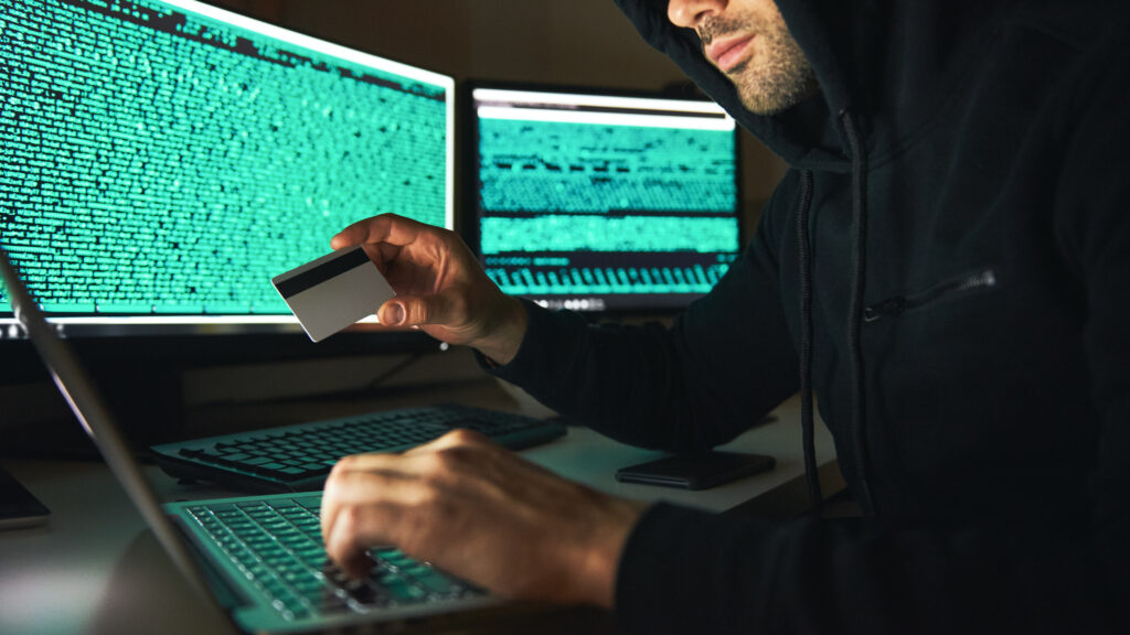 Stealing money. Side view of young bearded hacker in black hoodie using multiple computers and holding credit card while sitting in dark room. Binary code. Cyber attack. Cyber security